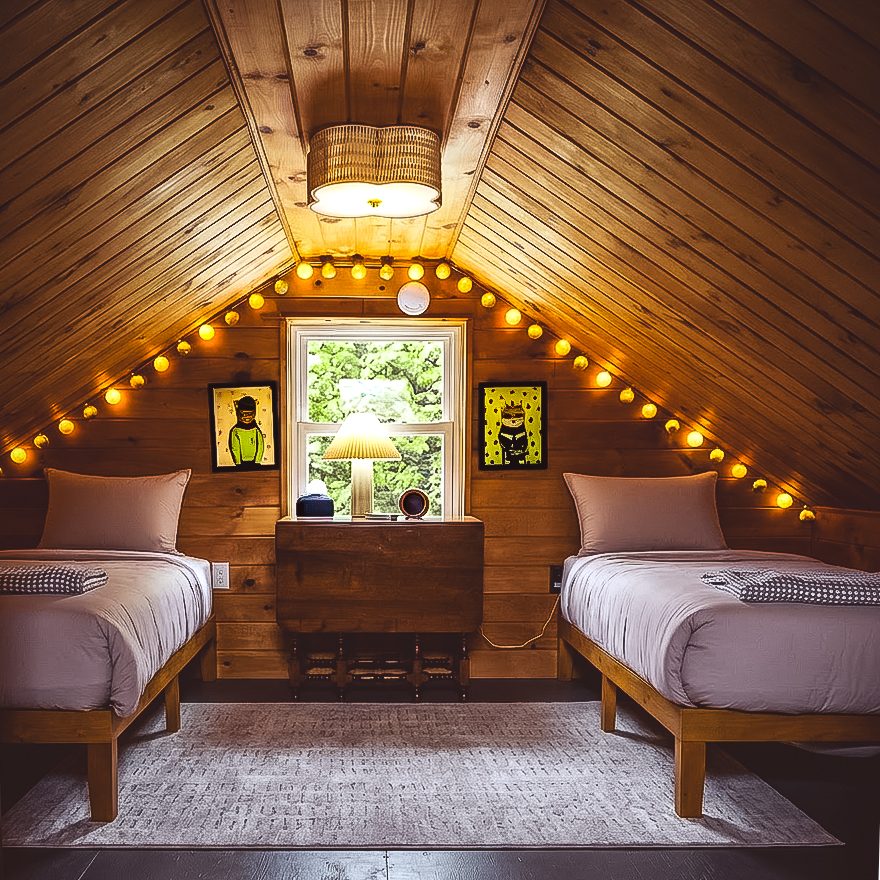 Two twin beds under the pitched wood ceiling of the Loft, strung with warm cafe lights, at a Catskills cabin near Livingston Manor, NY