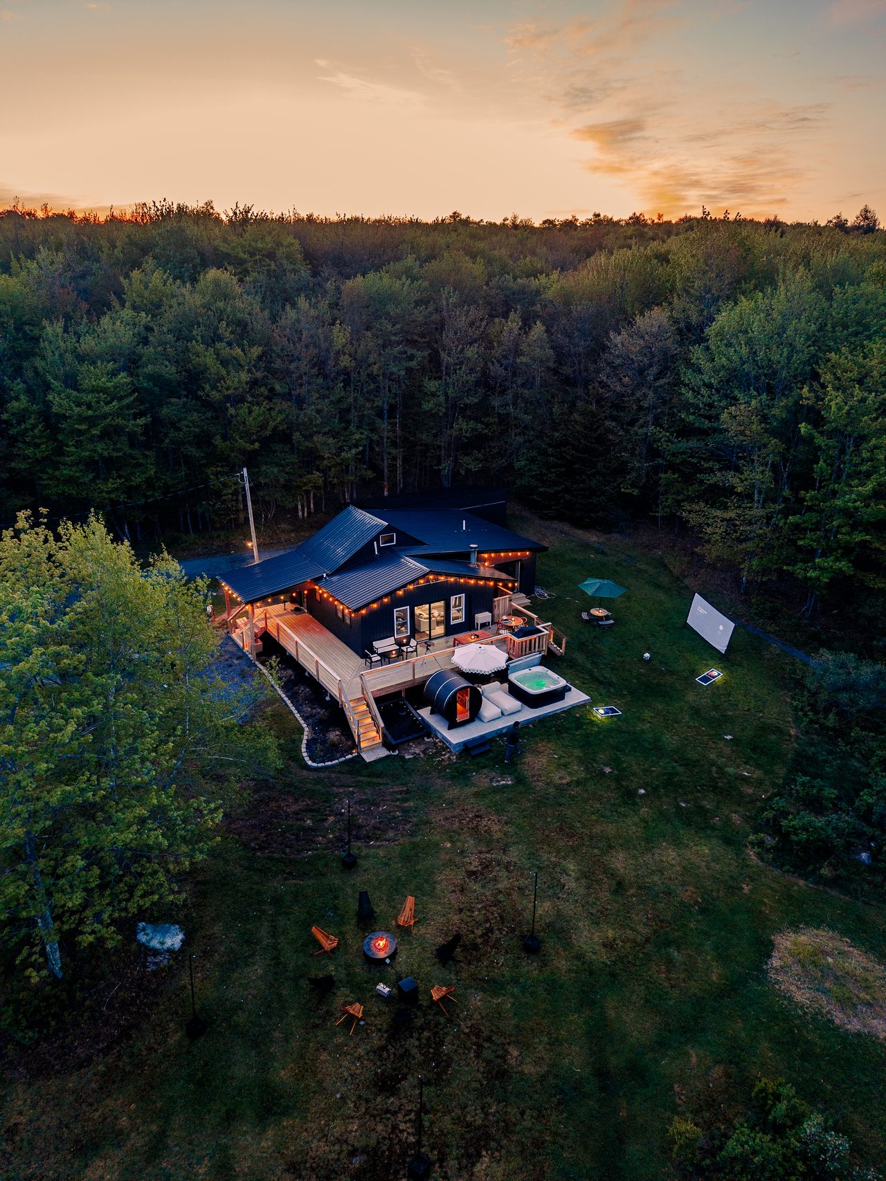 Aerial view at dusk of the cabin glowing with string lights, with friends gathered on the deck, a short drive from Bethel Woods