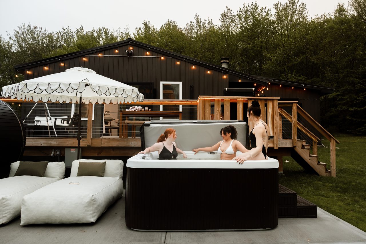 Friends chatting in the hot tub at dusk with the string-lit cabin, deck and barrel sauna behind, a western Catskills getaway near Livingston Manor, NY