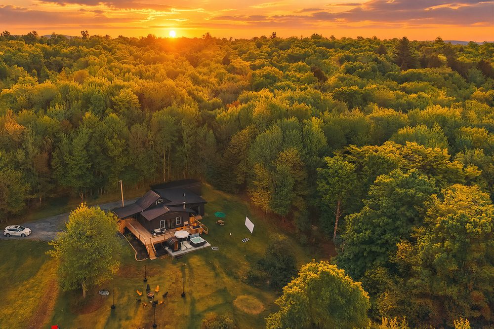 Aerial view at sunset of the black log cabin on five wooded acres, forest to the horizon in the western Catskills