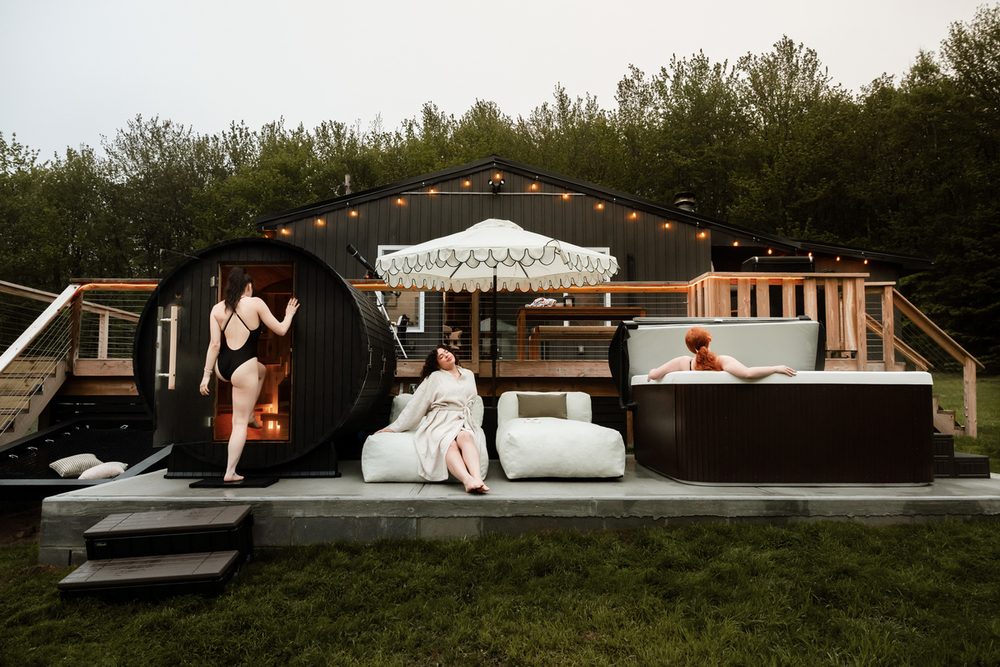 Barrel sauna and hot tub with lounge seating under string lights on the cabin's private deck