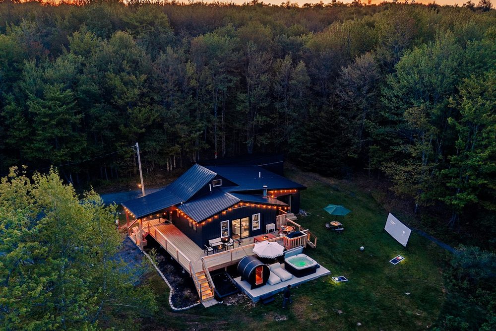 Aerial view at dusk of the string-lit cabin, deck, hot tub and barrel sauna in a forest clearing