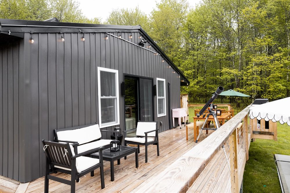 Black board-and-batten cabin exterior with deck seating, string lights and a telescope