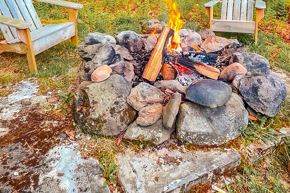 Stone-ringed wood-burning fire pit with Adirondack chairs in the woods