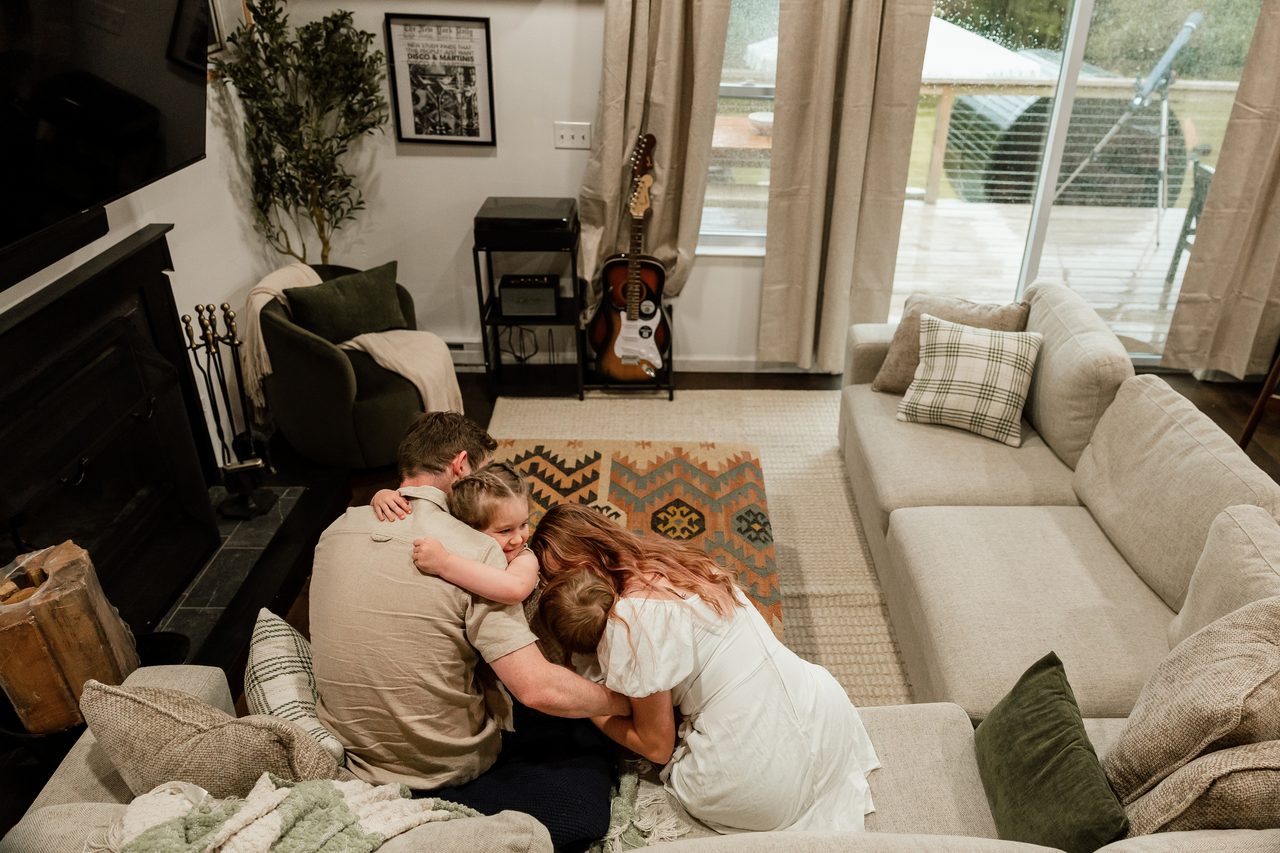 Family sharing a hug in the cabin's open living room with sectional sofas, a fireplace and deck views, Inkwood Reserve near Livingston Manor, NY